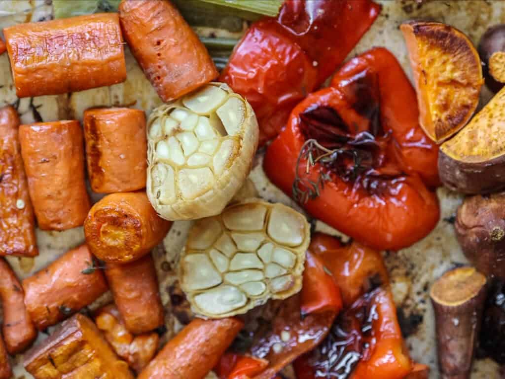 A close-up of roasted vegetables, including sliced carrots, red bell peppers, and garlic bulbs. The vegetables are caramelized and slightly charred, showcasing a rustic and appetizing presentation.